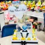 Father’s Day gift arrangement featuring a customized Father’s Day cake in a transparent cake box, hot air balloon decoration, yellow and white flowers, and premium display tray.