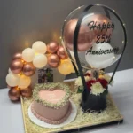 Luxury wedding anniversary gift arrangement featuring a heart-shaped anniversary cake, personalized hot air balloon decoration, illuminated balloon arch with couple photo, red and white floral arrangement, and premium wooden tray.